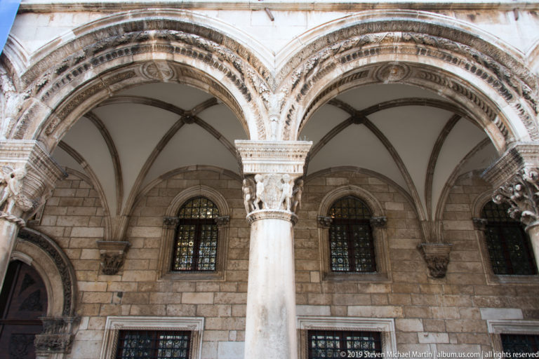 Vaulted stone arcade of the Rector’s Palace with carved capitals, ribbed ceilings, and barred windows behind.