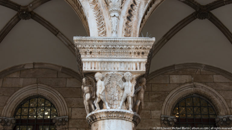Carved Rector’s Palace column capital with paired human figures, central shield, foliage, and layered ornamental molding.