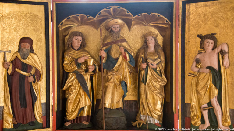 Gilded multi-panel altarpiece with four standing saints beneath carved arches against gold backgrounds.