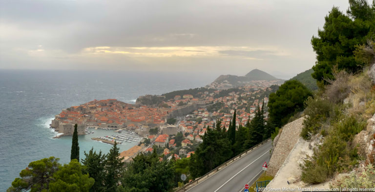 High hillside view of Dubrovnik’s walled Old Town beside the Adriatic under dramatic clouds.