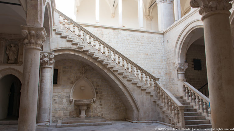 Monumental stone staircase with carved balustrade rising beneath an arch in the Rector’s Palace courtyard.