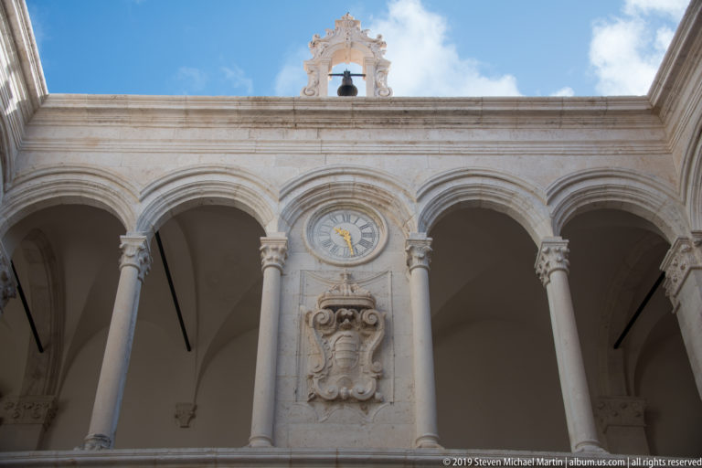 Upward view of Rector’s Palace courtyard arches with central clock, carved coat of arms, and small bell gable.
