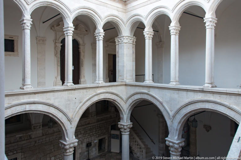 Corner of the Rector’s Palace courtyard with two levels of arches, slender columns, and a descending stone staircase.