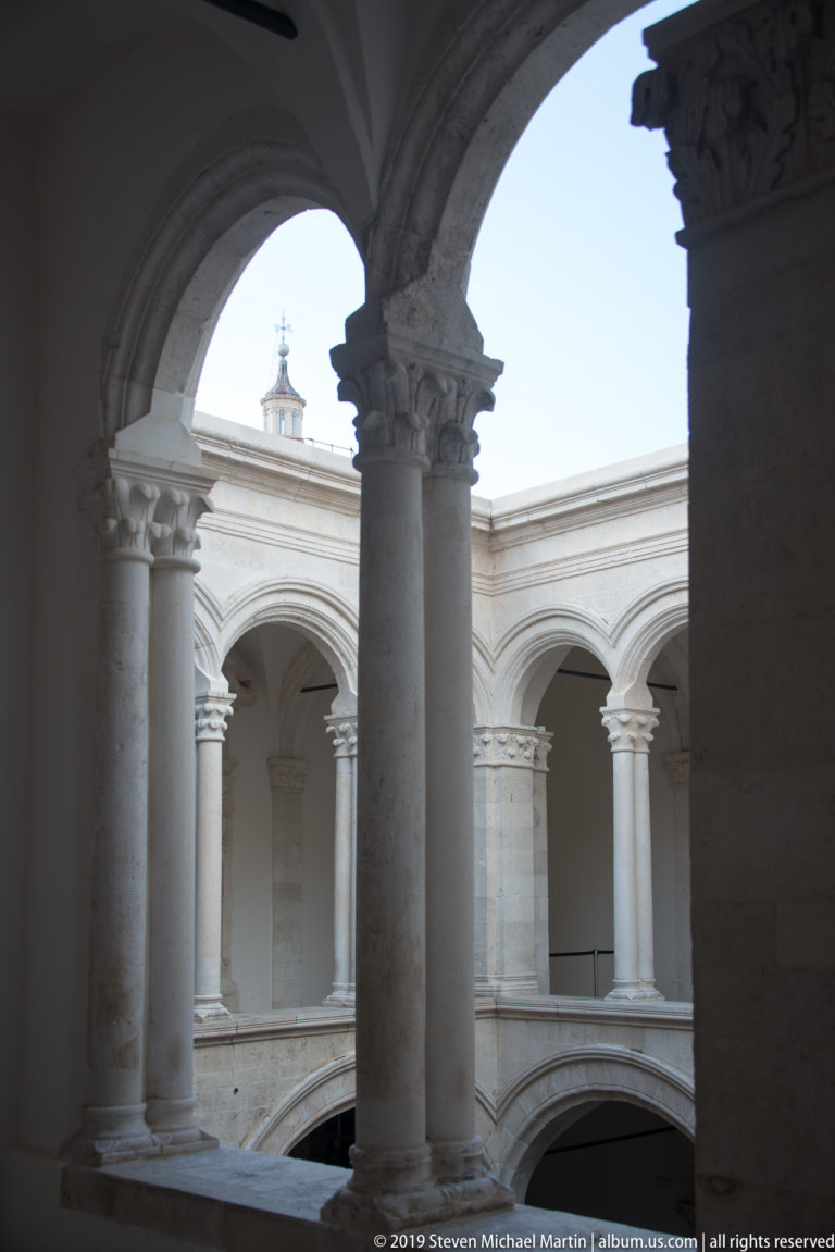 Dark stone arches and columns frame a sunlit upper arcade in the Rector’s Palace courtyard.