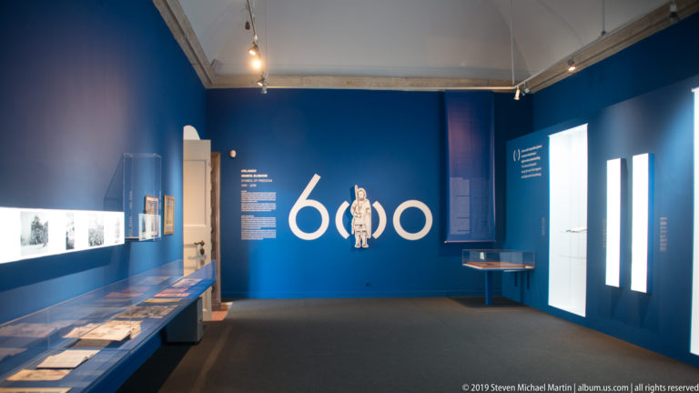 Blue museum gallery with a large white 600 graphic, lit display cases, documents, and interpretive wall text.