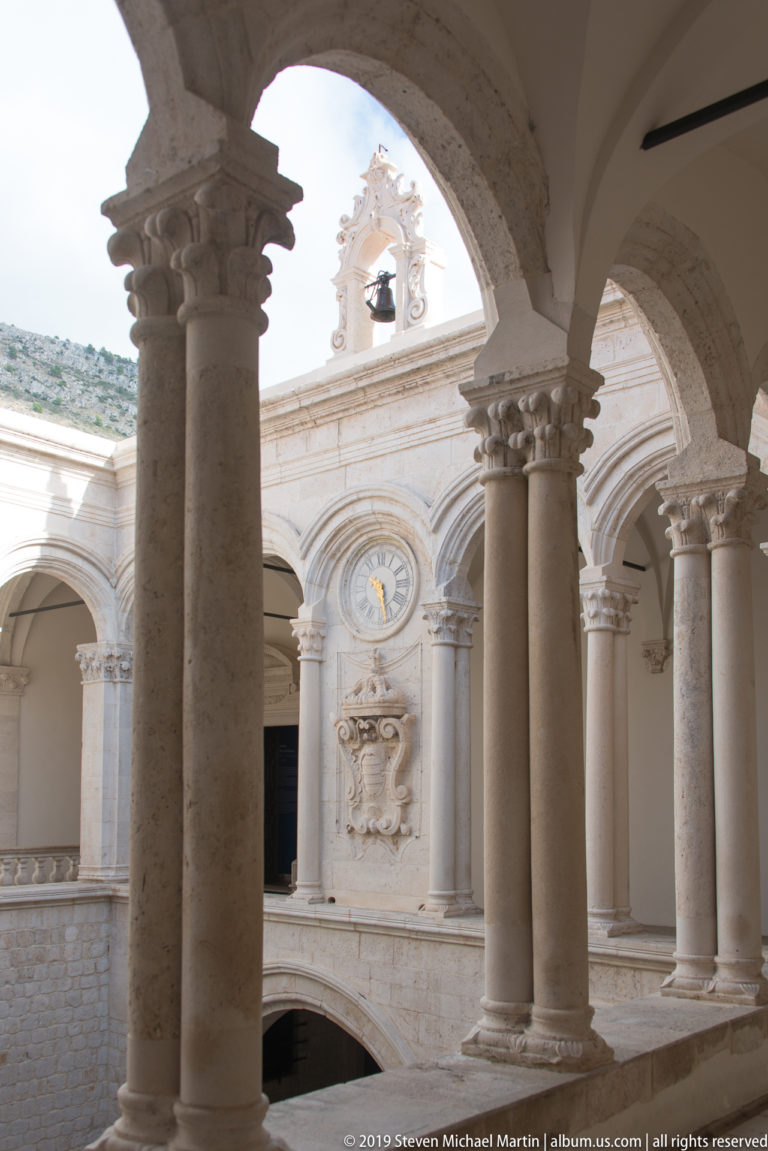 Stone arches frame the Rector’s Palace courtyard, with slender columns, upper arcade, clock, coat of arms, and bell gable.