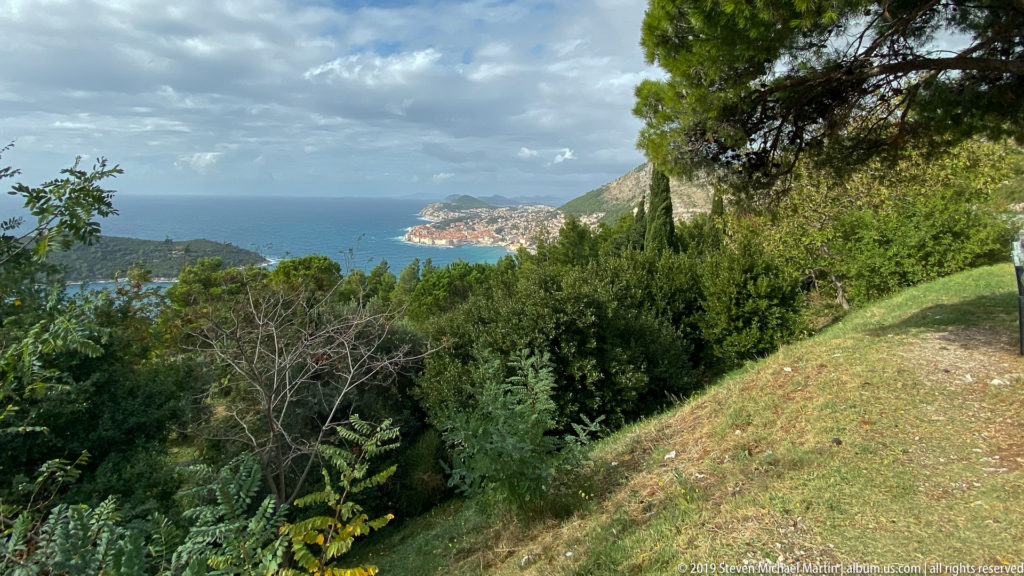 Grassy wooded hillside with shrubs and pines overlooking blue Adriatic water and a distant developed coastal headland.