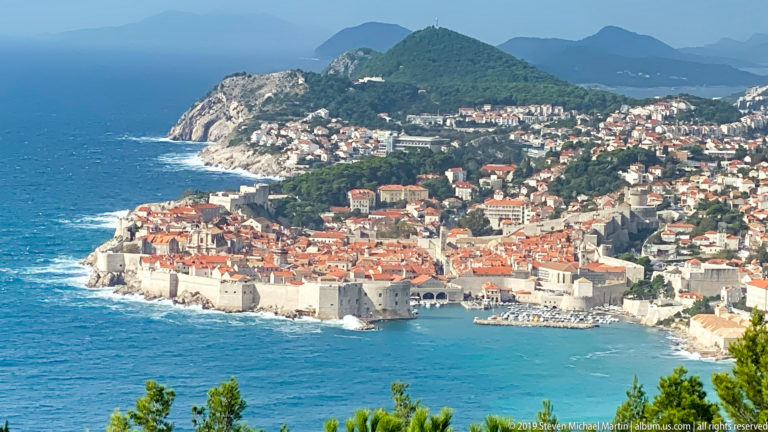 Panoramic elevated view of Dubrovnik Old Town, pale city walls, red roofs, blue Adriatic, and hillside neighborhoods.