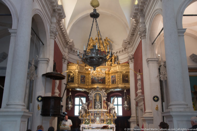 Church interior with gilded high altar, dark wooden pulpit, large hanging chandelier, and pale stone columns.