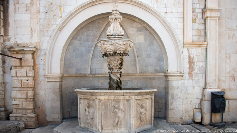Small Onofrio Fountain in an arched stone niche, with carved basin, twisted shaft, and ornate sculptural finial.