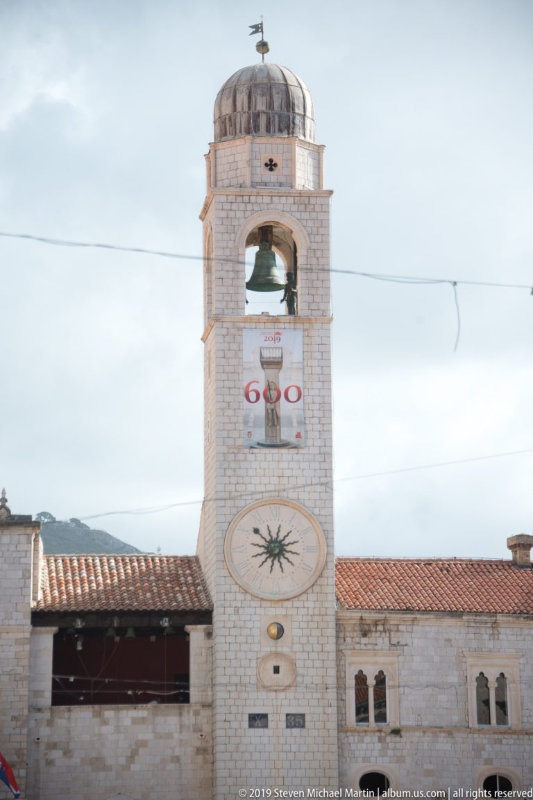 Dubrovnik City Bell Tower with civic clock, open bell stage, domed roof, and anniversary banner at Luža Square.