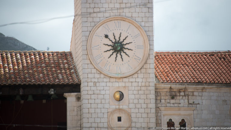 Clock face on Dubrovnik’s City Bell Tower with Roman numerals and the distinctive sun-shaped “Hobotnica” hand above red roof tiles.