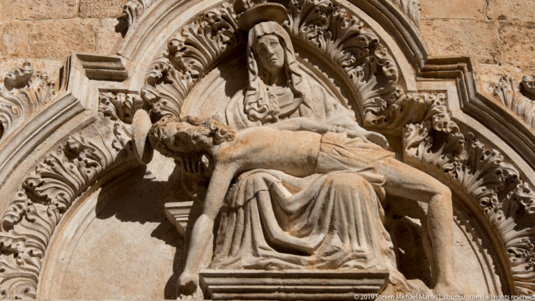Carved stone Pietà with the Virgin Mary holding Christ beneath an ornate foliage-filled architectural canopy.