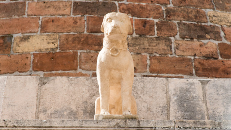 Small weathered pale stone dog or hound sculpture standing on a ledge against a red brick wall.