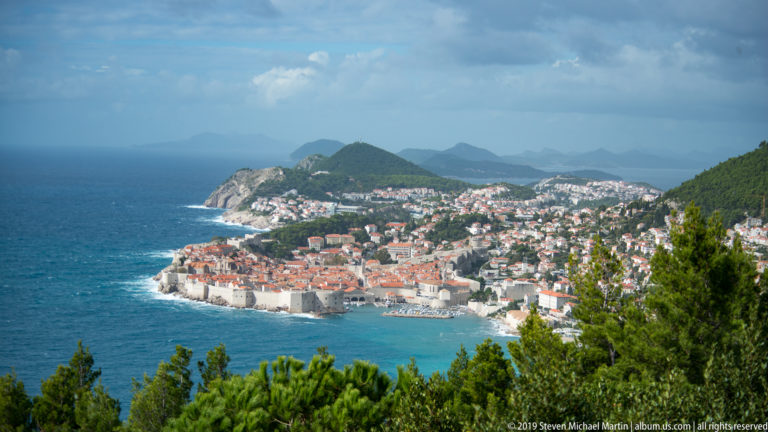 Distant view through pine trees toward Dubrovnik Old Town, its red roofs and pale walls beside the blue Adriatic.