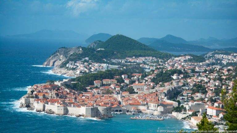 Elevated panorama of walled Dubrovnik beside the Adriatic, with modern neighborhoods spreading across green hills beyond.