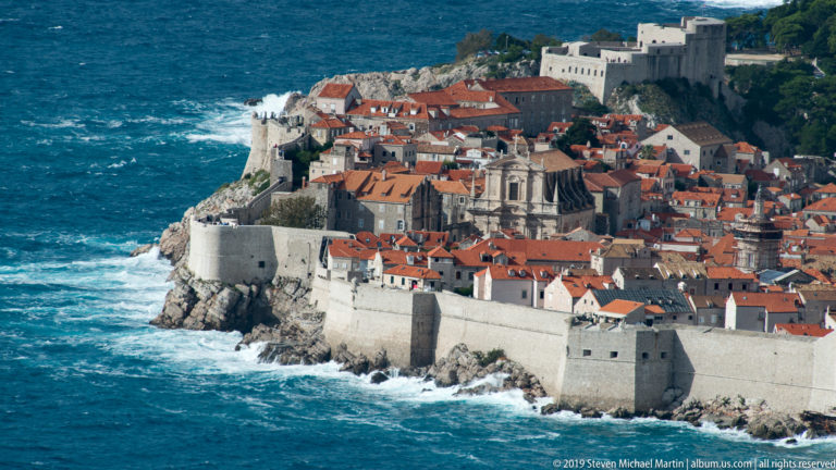Dubrovnik Old Town enclosed by pale stone sea walls above the Adriatic, with red roofs and church domes inside.