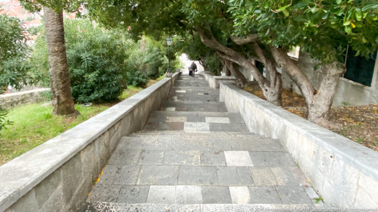Long stone stairway climbing beneath mature trees and bordered by low pale walls.