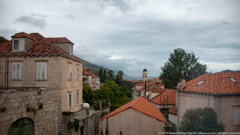 Stone houses with red-tiled roofs descending toward Dubrovnik, with a distant tower and mountain ridge.