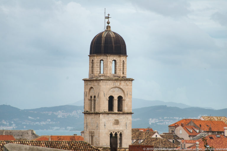 Tall pale-stone bell tower with arched openings and a dark dome rising above Dubrovnik’s red-tiled roofs.