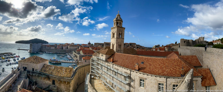 Stone bell tower above Dubrovnik rooftops, with harbor, Adriatic water and Lokrum behind.