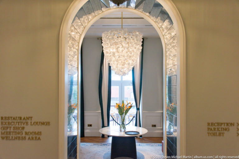 Arched hotel interior with chandelier, round table, tall windows and directional wall lettering.