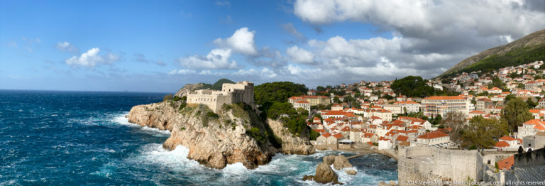 Fortress on rocky coast beside Dubrovnik walls, red roofs and rough Adriatic water.