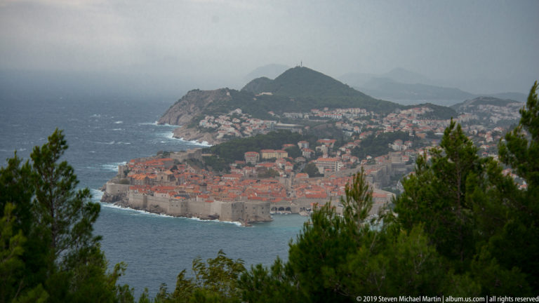 Distant high view of Dubrovnik’s walled red-roofed Old Town with wooded hills and coastal neighborhoods.