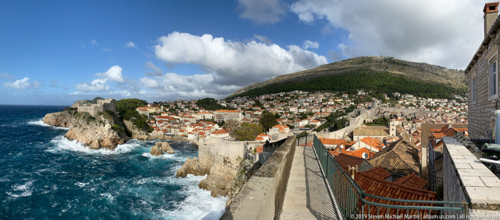 Wide view of Dubrovnik walls and red roofs between rough Adriatic sea and green hillside.