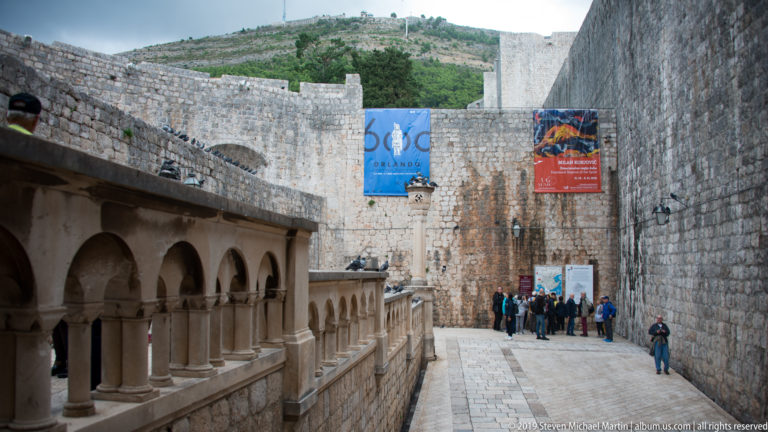 Visitors walking through a broad stone passage between high Dubrovnik fortification walls.
