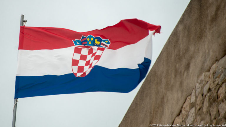 Croatian red, white and blue flag with checkerboard coat of arms blowing beside stone wall.