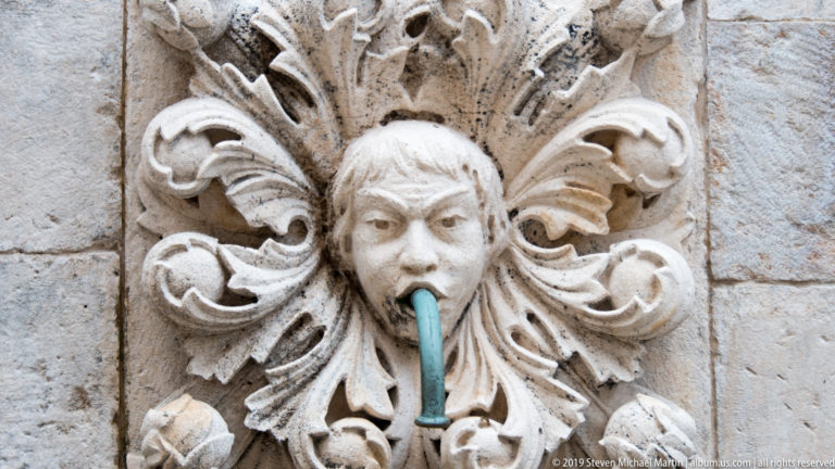 Carved stone human face with metal water spout surrounded by ornate foliage.