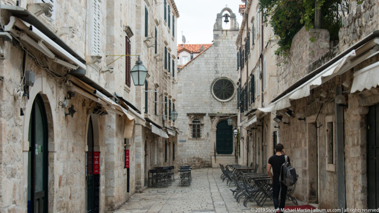 Narrow stone street with café seating leading toward a small church façade.
