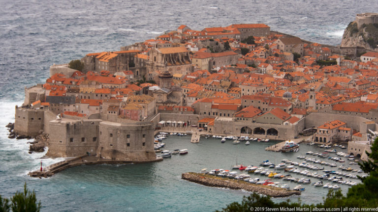 Elevated view of Dubrovnik’s coastal fortress, small marina and red-roofed Old Town beside the sea.