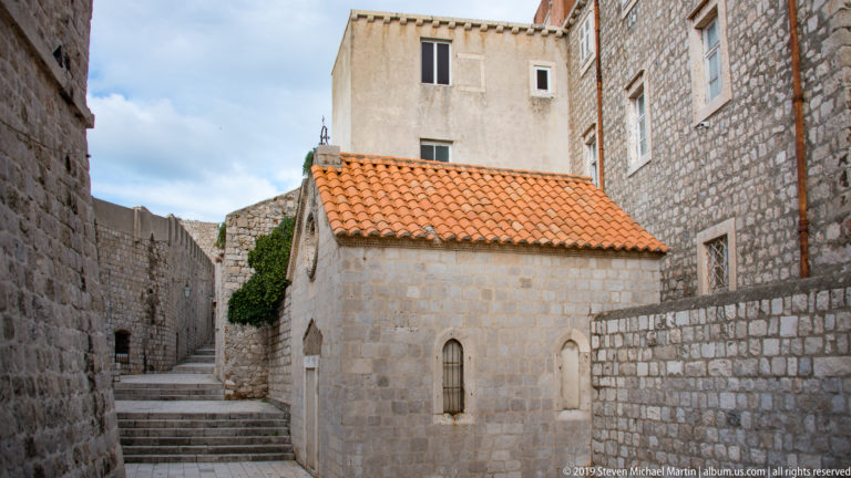 Small stone chapel with red-tile roof and narrow arched windows beside stepped lanes.