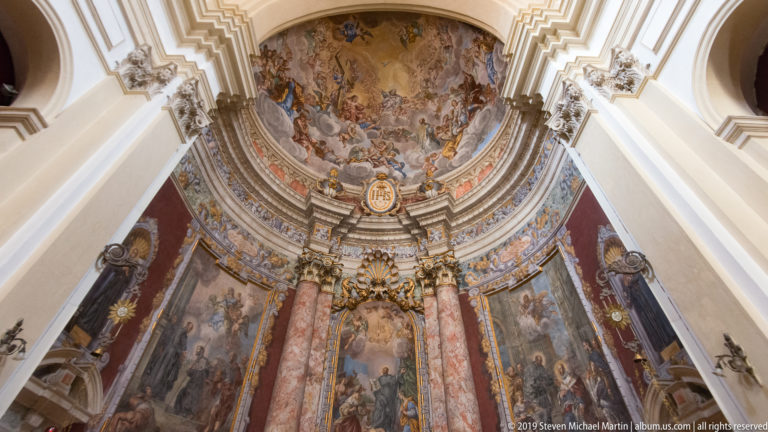 Upward view of a Baroque altar with red marble columns beneath a large painted ceiling fresco.