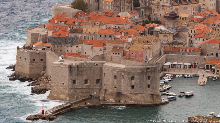 Large stone waterfront fortress below Dubrovnik’s dense red-roofed Old Town, with small boats alongside.