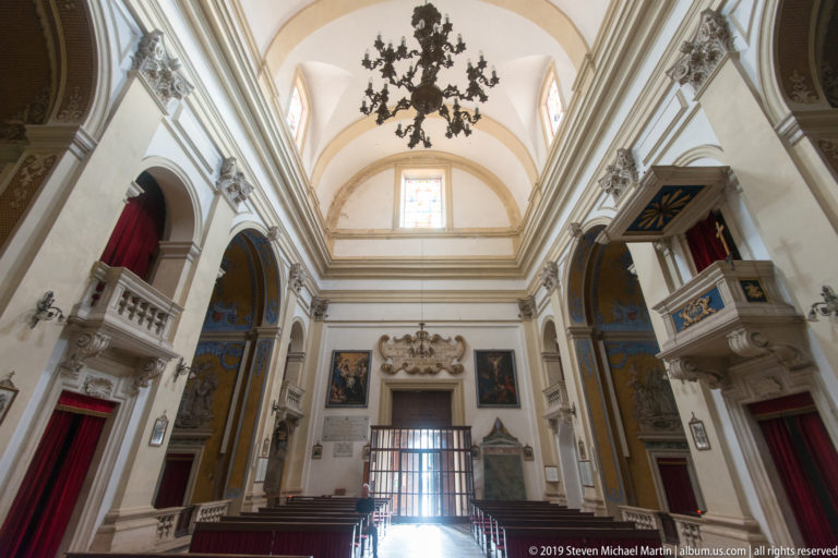Baroque church nave with side chapels, pulpit, dark chandelier and high white vaulted ceiling.