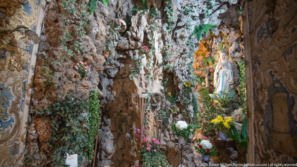 Statue of the Virgin Mary inside a rocky grotto shrine decorated with flowers and greenery.