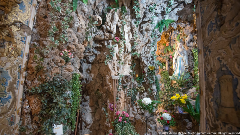 Statue of the Virgin Mary inside a rocky grotto shrine decorated with flowers and greenery.