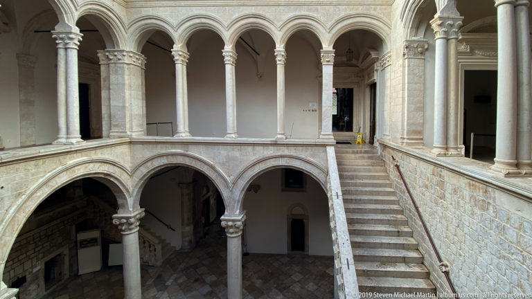 Two-level stone courtyard with arcades, slender columns and a staircase between galleries.