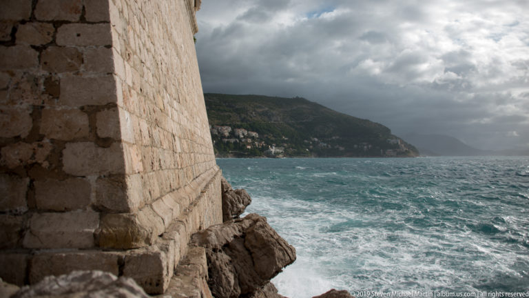 Tall stone city wall beside rough turquoise sea and rocky shoreline under dark clouds.