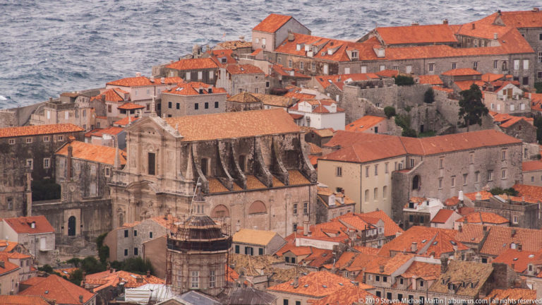 Dense Dubrovnik terracotta rooftops surrounding large pale-stone churches and historic buildings.