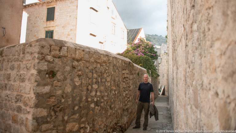 Man walking along a narrow stone lane between high masonry walls in Dubrovnik.