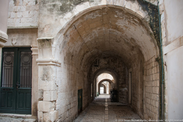 Long stone passage with repeated rounded arches, worn paving and dark doorways.