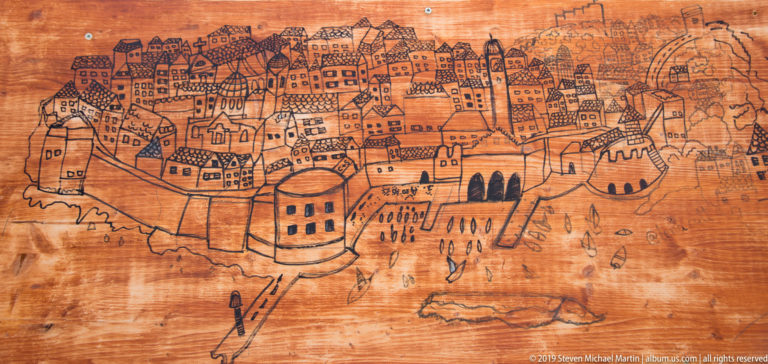 Line drawing of Dubrovnik’s walled city, towers and harbor defenses on a wooden board.