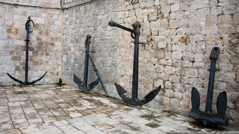 Four large historic iron anchors displayed upright and leaning against a stone wall.