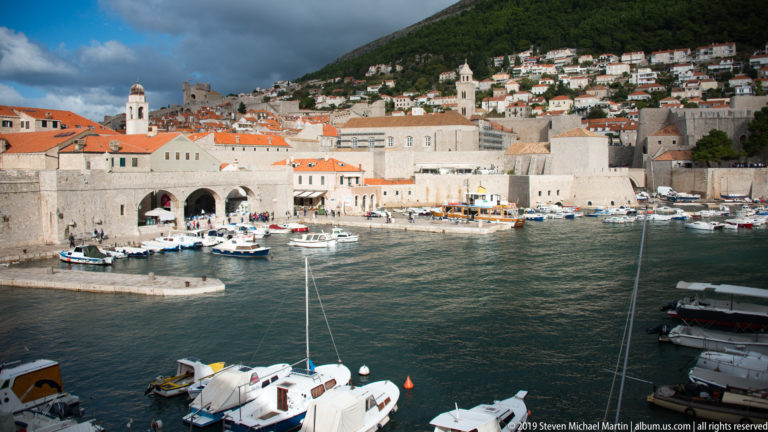Wide view of Dubrovnik Old Port with boats, stone walls, red roofs and hillside houses.