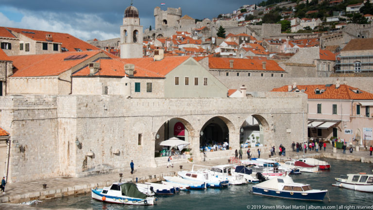 Boats moored in Dubrovnik’s Old Port below broad stone arches and red-roofed Old Town buildings.