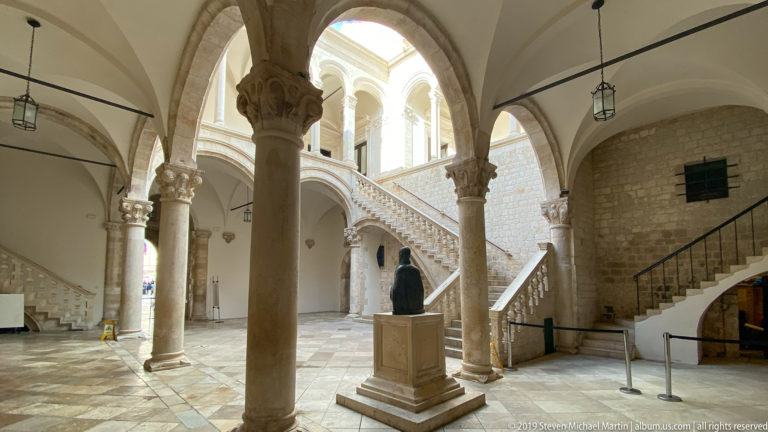 Two-story pale stone courtyard with arcades, columns and an open staircase.